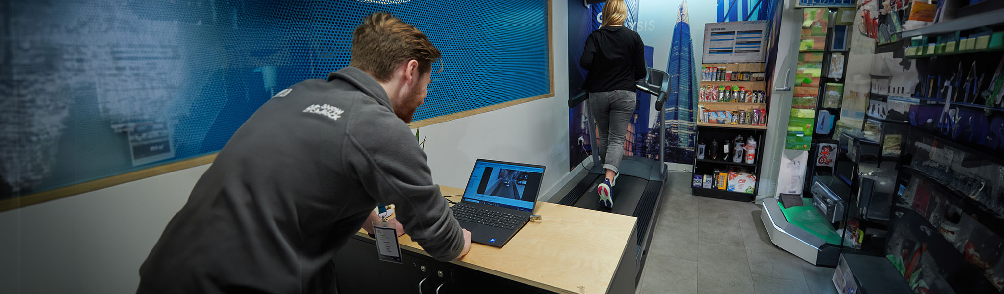 A man in a shop is performing a gait analysis for a customer on a treadmill