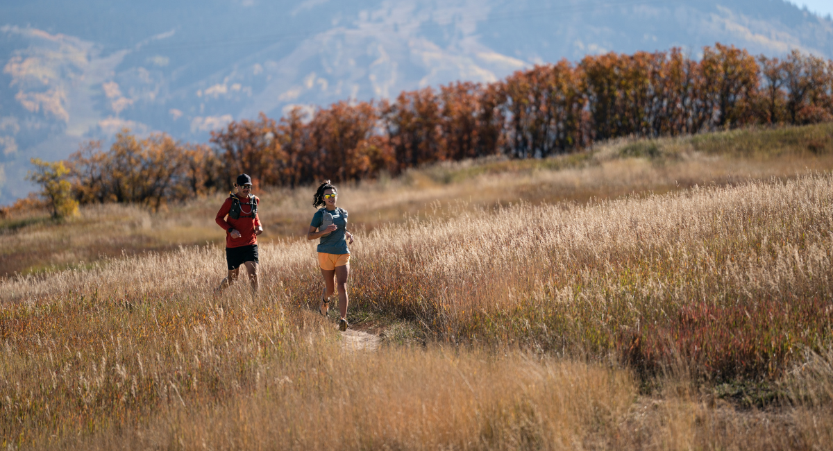 A woman and a man run through a field of wheat in the sunshine with golden, autumn coloured trees and the mountains behind them