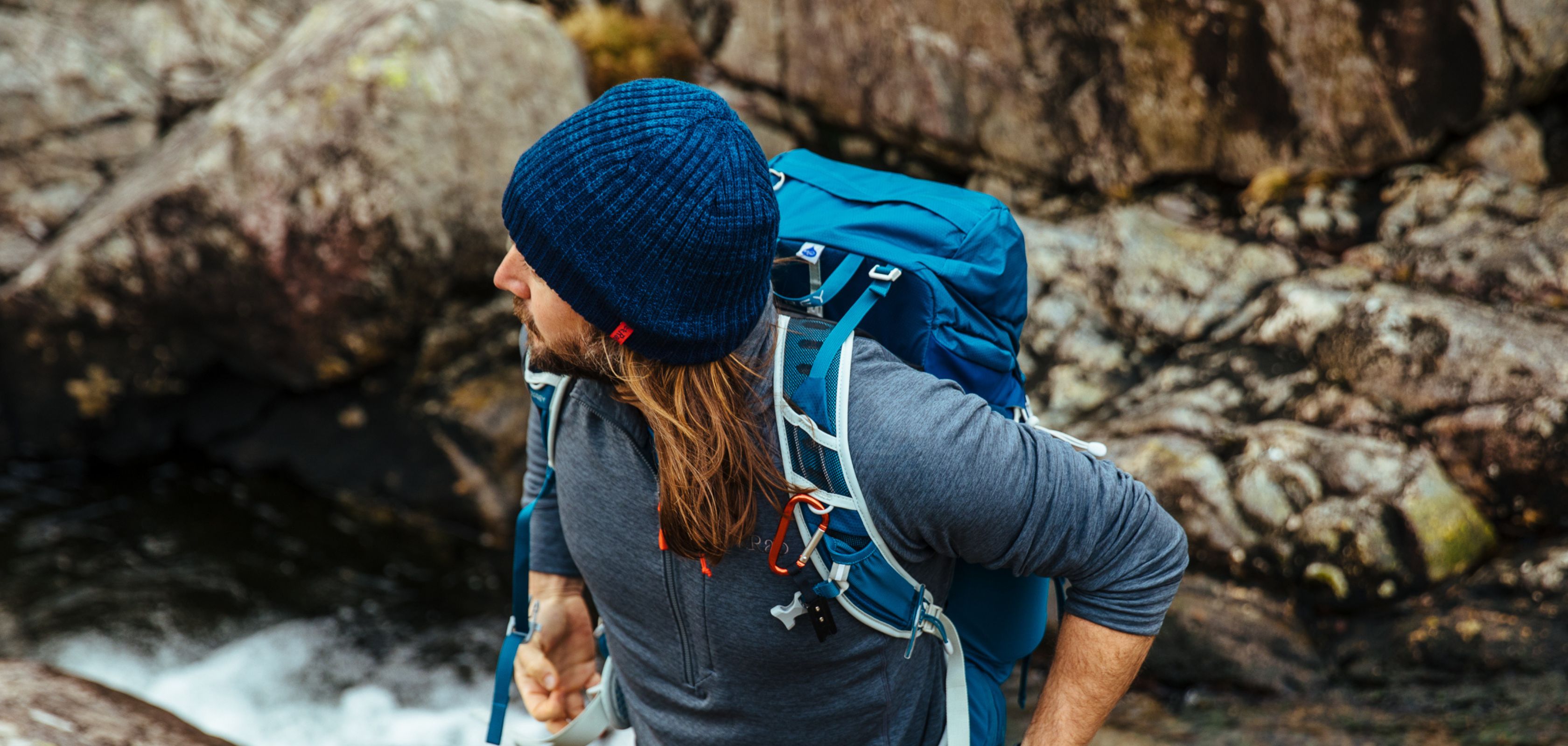 A man in a blue wooly had and rucksack climbing a rugged mountain