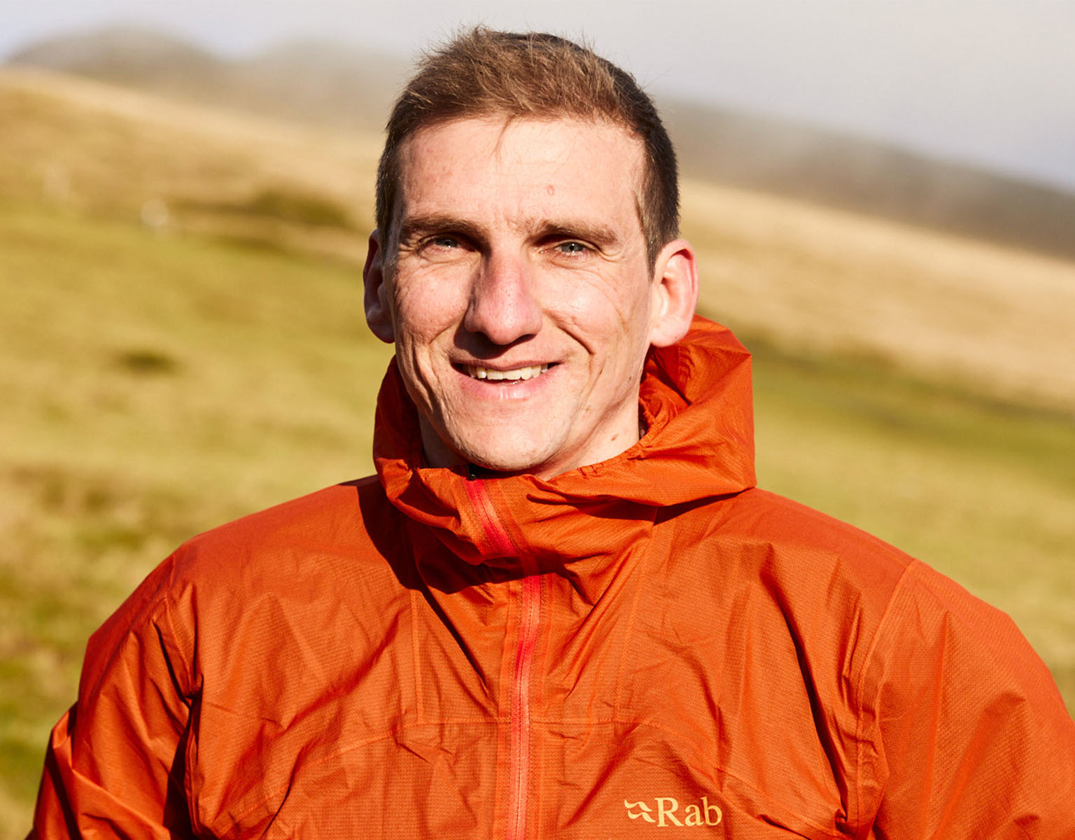 A man is smiling somewhere outside, wearing an orange Rab waterproof jacket, with a yellow grass fieald behind him